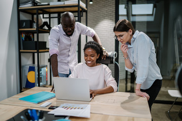 Wall Mural - Cheerful afro american female director showing funny video content to professional colleagues during work break in conference room, successful diverse entrepreneurs enjoying chatting via netbook