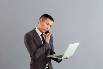 Wall Mural - Asian businessman with laptop talking by phone on grey background