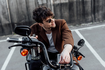 young man in sunglasses holding bottle, sitting on motorcycle and looking away