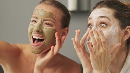 two pretty smiling girls friends with facial mask on their faces posing and enjoying the moment at h