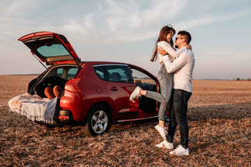 Wall Mural - Young Happy Couple Dressed Alike in White Shirt and Jeans Enjoying Road Trip at Their New Car, Beautiful Sunset on the Field, Vacation and Travel Concept