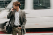 © Anton - Careful young businessman walking on crosswalk and look to left. Talk on phone. Car riding behind. Blurred background. Posing on camera. Hold purse on shoulder.