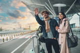 Fototapeta  - Young man and woman stand outside airport and wait for taxi cab. Guy wave with hand. After vacation or travel. Georgian woman hold his hand and smile. Sunset outside. Evening.