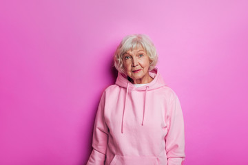 Wall Mural - Amazing old woman in pink sweater looks modern and good-looking. Stand alone and look forward. Calm, peaceful and confident senior female model in hoody. Isolated over pink background.