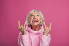 Emotional, Cheerful And Happy Old Woman In Modern Pink Hoody Posing On Camera Alone. Look Up And Laughing. Lady With Grey Hair Show Rock Signs. Isolated Over Pink Background.