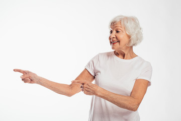 Wall Mural - Happy cheerful old woman with grey hair point with fingers on left and smile. Stand alone and pose on camera. Move and action. Have fun alone. Isolated over white background.