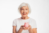 Picture of old woman with grey hair holding pink jar of face cream in hands. Look straight and smile. Anti age care and treatment at home. Isolated over white background.