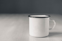 Enamel White Metal Mug With Black Line On The Edge On White Wooden Table On Gray Background. Place For Text Or Advertising