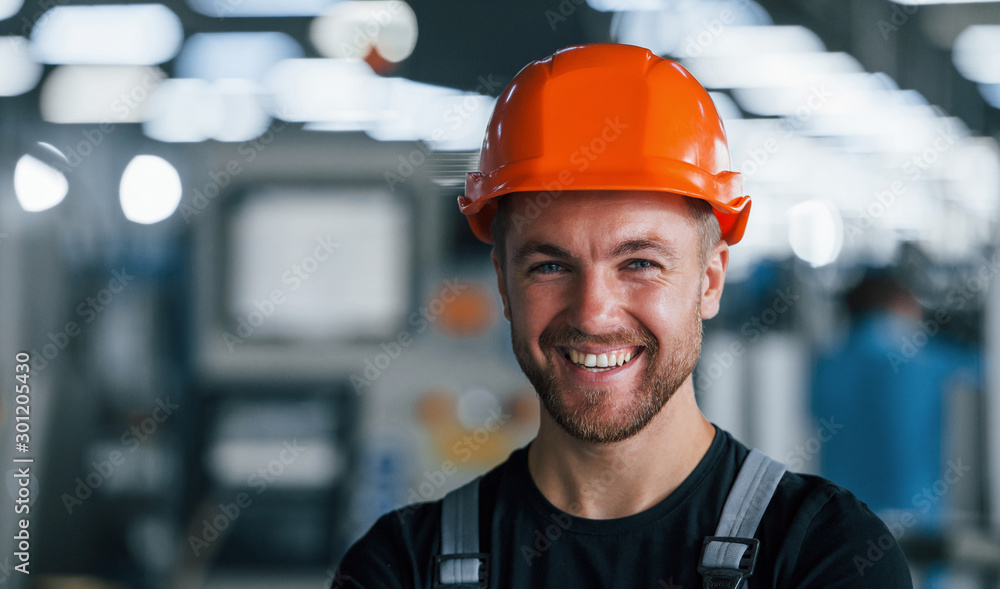 Smiling and happy employee. Portrait of industrial worker indoors in factory. Young technician with orange hard hat - obrazy, fototapety, plakaty 
