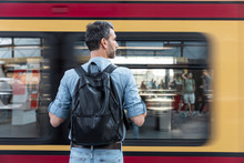Rear View Of Man With Backpack At The Station Platform While Train Coming In, Berlin, Germany