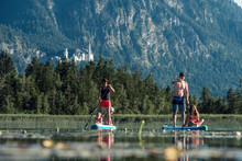 Family Paddle Boarding On Lake Bannwaldsee, Castle Neuschwanstein In The, Fuessen, Germany
