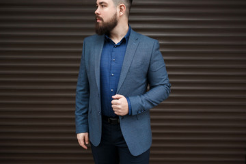 Stylish handsome brunette man with beard, wearing suit jacket and blue shirt, outdoors on the city street near brown roller door. Details of classic elegant formal men's outfit. Businessman.