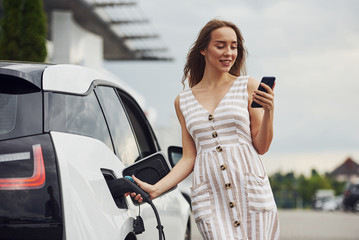 Using smartphone while waiting. Woman on the electric cars charge station at daytime. Brand new vehicle