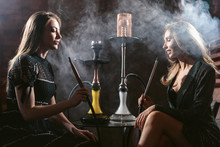 Girls Party In Hookah Lounge. Group Of Two Young Women In Backlight Smoking Shisha In Cafe Or Bar, Making Smoke Clouds, Having Fun, Smiling. Relax Concept. Friendship