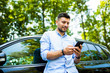 © F8  \ Suport Ukraine - Checking social media. Young attractive smiling man with beard standing near his car and holding mobile phone