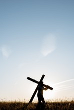 Vertical Shot Of A Male Carrying A Handmade Wooden Cross In A Grassy Field Under A Blue Sky