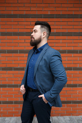 Wall Mural - Stylish handsome brunette man with beard, wearing blue suit jacket, shirt and pants, standing outdoors on the city street near red brick wall. Classic elegant formal men's outfit. Businessman portrait