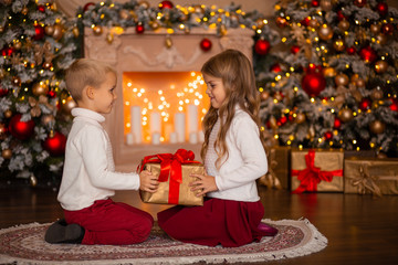 Wall Mural - in anticipation of the New Year holiday. Twins boy and girl are sitting near the fireplace on the carpet against the background of Christmas trees with lights. They dream of something and give each ot