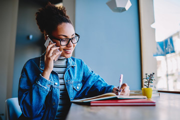 Wall Mural - Happy female student calling to professor from university for discussing details about upcoming exam notes main theses in textbook for education, positive hipster girl making telephone conversation