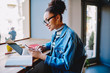 © BullRun - Positive african hipster girl in spectacles messaging via cellphone during e learning on laptop computer, happy smiling woman search contact of colleague for dialing number via smartphone device