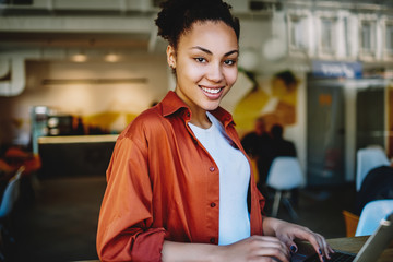 Wall Mural - Portrait of african skilled woman smiling and looking at camera in break of working remotely on laptop coputer, happy dark skinned hipster girl enjoying free high speed internet in cafeteria