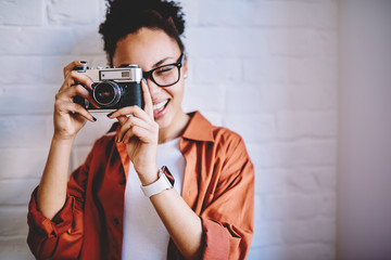 Close up portrait of cheerful afro american female photographer clicking images via retro camera during free time, happy dark skinned amateur spending time for favourite hobby with vintage equipment