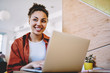 © BullRun - Cheerful hipster girl sitting with modern netbook in coworking space and laughing from received funny message, positive black woman looking away and smiling during e learning in cafe interior