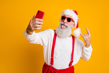 X-mas party hard. Portrait of funny crazy santa claus in red hat take selfie smart phone christmas blogger show horns grimace tongue out wear shirt suspenders isolated yellow color background