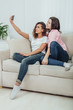 © Andrii - Cute young girlfriends sitting on the sofa, taking selfie, posing, making faces.
