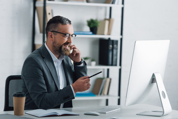 Wall Mural - handsome businessman in formal wear talking on smartphone and pointing at computer