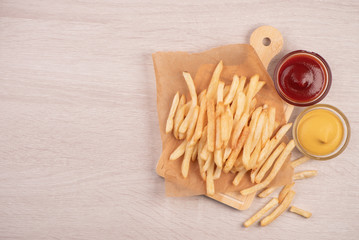 Golden yummy deep French fries on kraft baking sheet paper and serving tray to eat with ketchup and yellow mustard, top view, lifestyle.