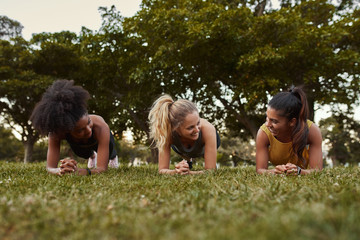 Canvas Print - Three sporty young women smiling while doing plank exercises lying on green grass in the park