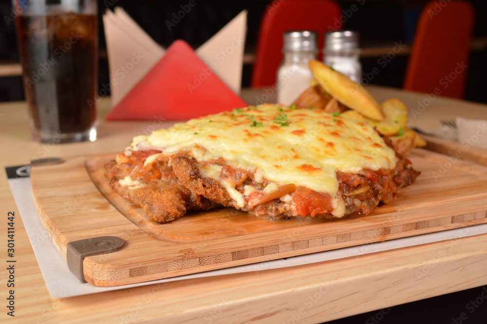 CLASSIC BREADED FRIED MEAT , KNOWN IN ARGENTINA AS MILANESA, CHEESE AND TOMATO, SERVED WITH FRENCH FRIES ON A WOODEN TABLE
