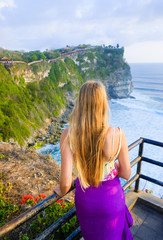 Wall Mural - Blonde girl with long hair in traditional purple skirt at sunset on ocean background and Uluwatu temple, Bali, Indonesia