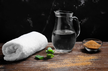 White terry towel, cane sugar, aloe and a jug of water on a natural wood table.