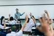 © pop_thailand - An smiling Asian male high school teacher teaches the white uniform students in the classroom by asking questions and then the students raise their hands for answers.