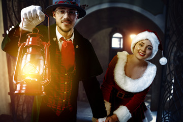 Poster - Two friends inside. Friends walking down the corridor with a lighted lamp. A young woman in a red dress and Santa's hat. A man in a tuxedo and a gray hat with a red lamp.