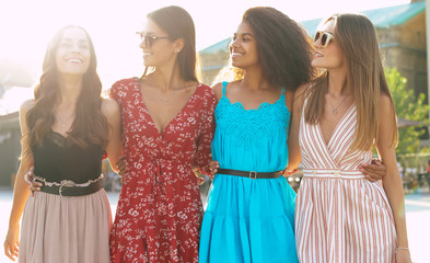 Wall Mural - Female Gracefulness. Four attractive female friends in bright dresses of different colors wearing sunglasses are hugging each other by waists, smiling broadly and looking at each other.