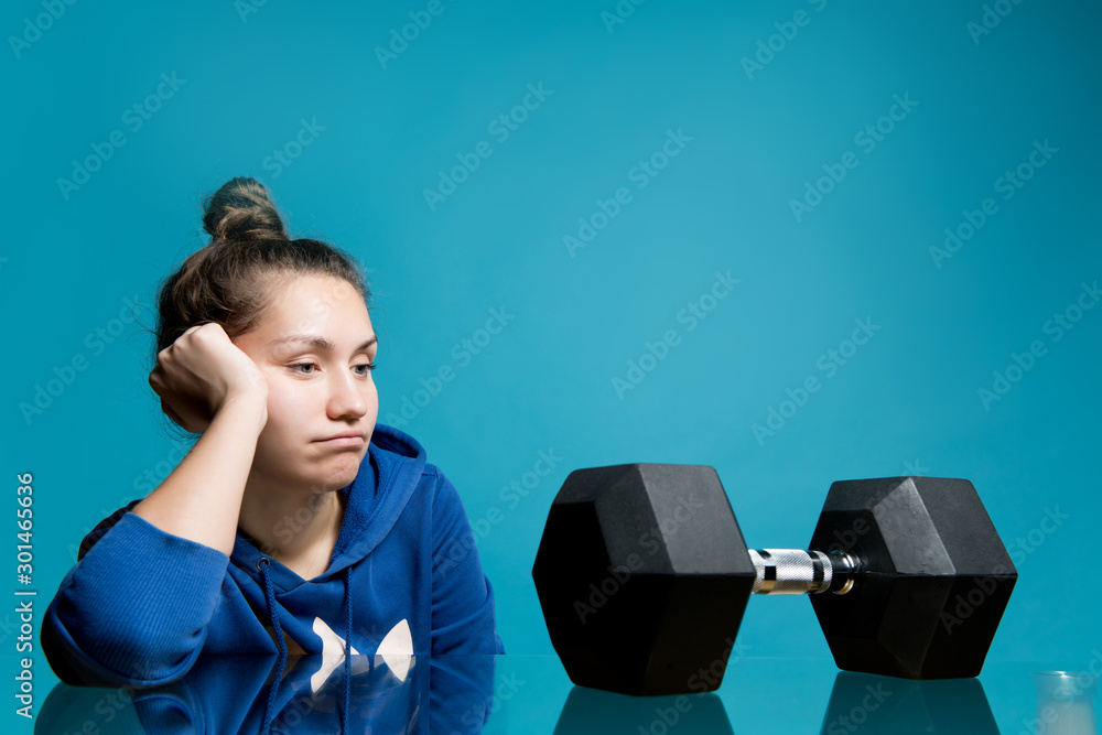 the girl looks in frustration and longing at the big dumbbell lying in front of her - obrazy, fototapety, plakaty 