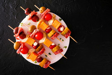 Skewered Party Appetizers With Meat, Cheeses And Pickles On A Serving Plate. Overhead View On A Dark Stone Background.