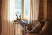 Young Man In Warm Sweater Reading By The Window Inside Cozy Log Cabin