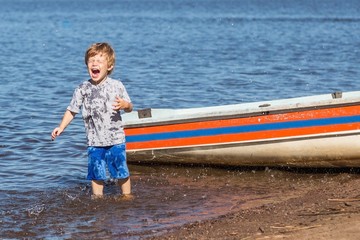 little cute redhead Caucasian boy enjoying splashes of water near boat in the lake playing funny game with parents in summer sunny day