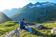 A Young Man Cooking A Dinner In The Wilderness During The Sunrise. He Is Sitting At The Edge Of A Mountain, Mixing The Dish. He Is Surrounded By Tall Mountains. Alpine Camping. Outdoor Kitchen.