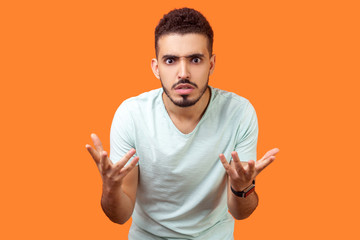 What do you want? Portrait of disgruntled annoyed brunette man with beard in casual white t-shirt standing with raised hands, mad indignant expression. indoor studio shot isolated on orange background