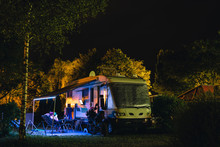 People Sit Under An Awning And Watch TV, Night View Of The Parking Lot For A Motorhome, Camper Van, Campsite Camp For Sleeping And Relaxing. Vacation And Travel Tour. Motorcycle.