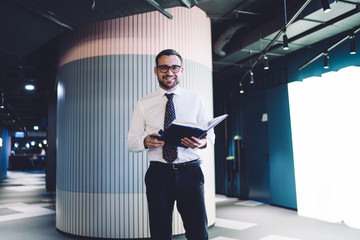 Portrait of man successful bookkeeper holding textbook with accounting information and smiling at camera during working day, happy male in optical spectacles standing with notepad for notary