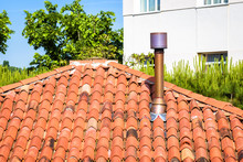 Chimney On Roof Of Residential House. Home Round Chimney Close-up In Summer. Modern Air Vent System On Rooftop. Metal Chimney Pipe On Background Of Red Tiles, Trees And Building.