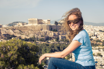 Wall Mural - Young pretty woman sits at hilltop in Athens, Greece. Beautiful adult girl tourist smiles overlooking Athens city center. Attractive happy person against famous Acropolis. Concept of travel in Athens.