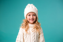 Little Blonde Girl In White Winter Knitted Hat And Sweater On Blue Background Isolate, Space For Text. The Child Rejoices In Winter And Snow. Winter Clothes