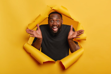 Wall Mural - Merry black man spreads palms, has joyous expression, white teeth, makes decision, gets unexpected offer, wears dark clothing, stands in paper hole, gestures from happiness, glad to be praised